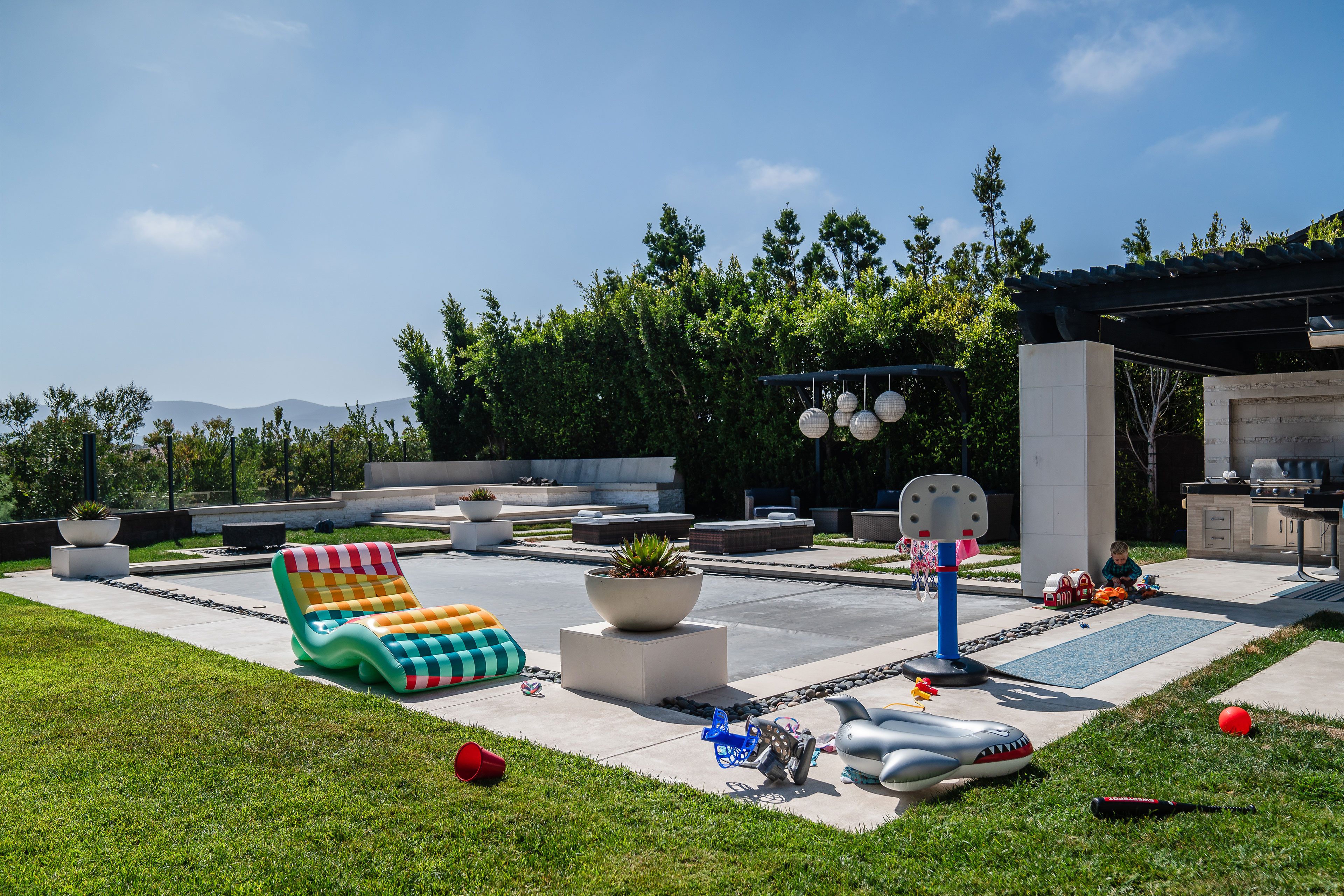 A fenced-in backyard shows a covered pool, firepit with seating, a pool chair, and toys.