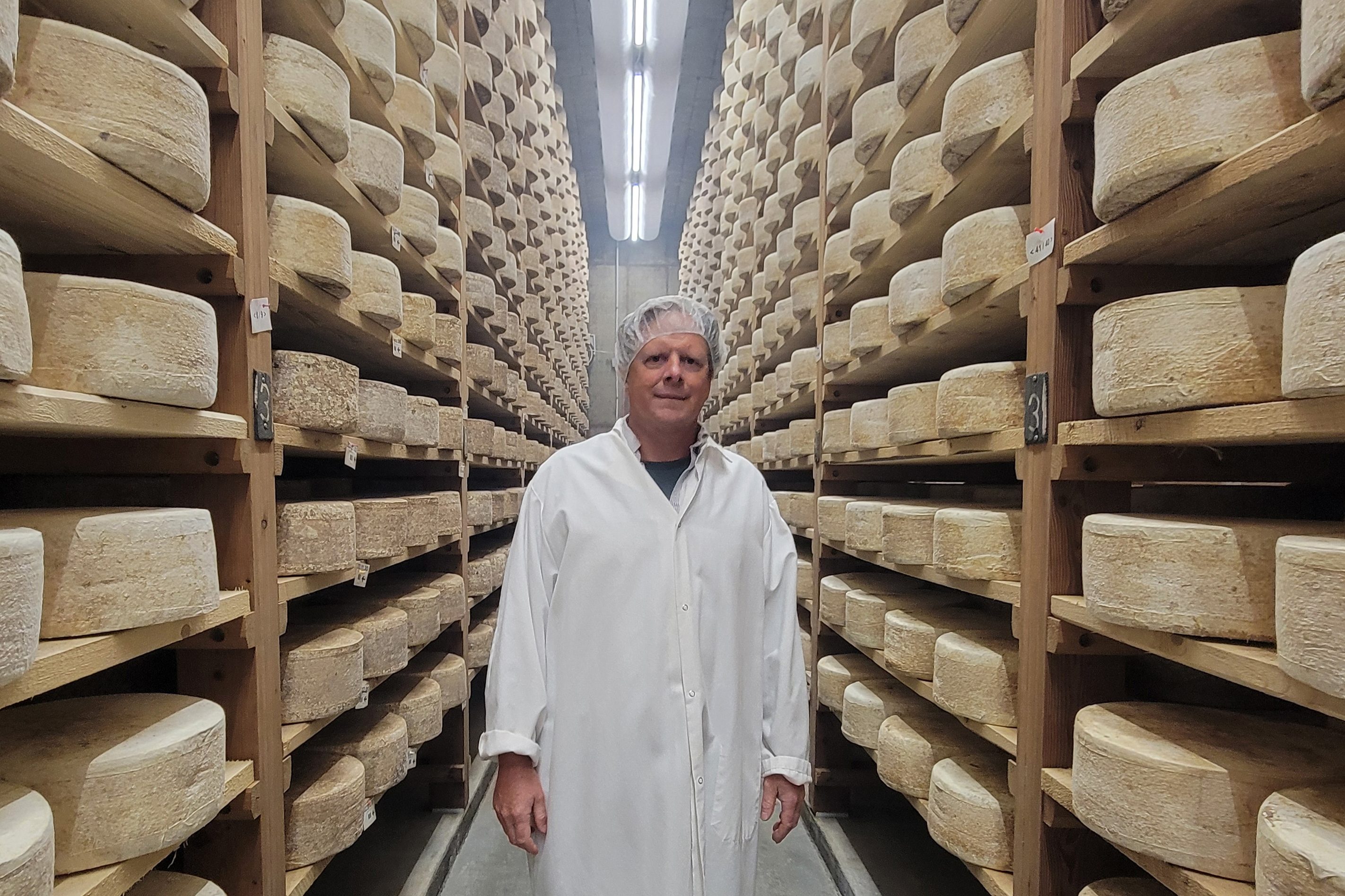 A photo of a man standing in a room surrounded by shelves of ripening cheese wheels.