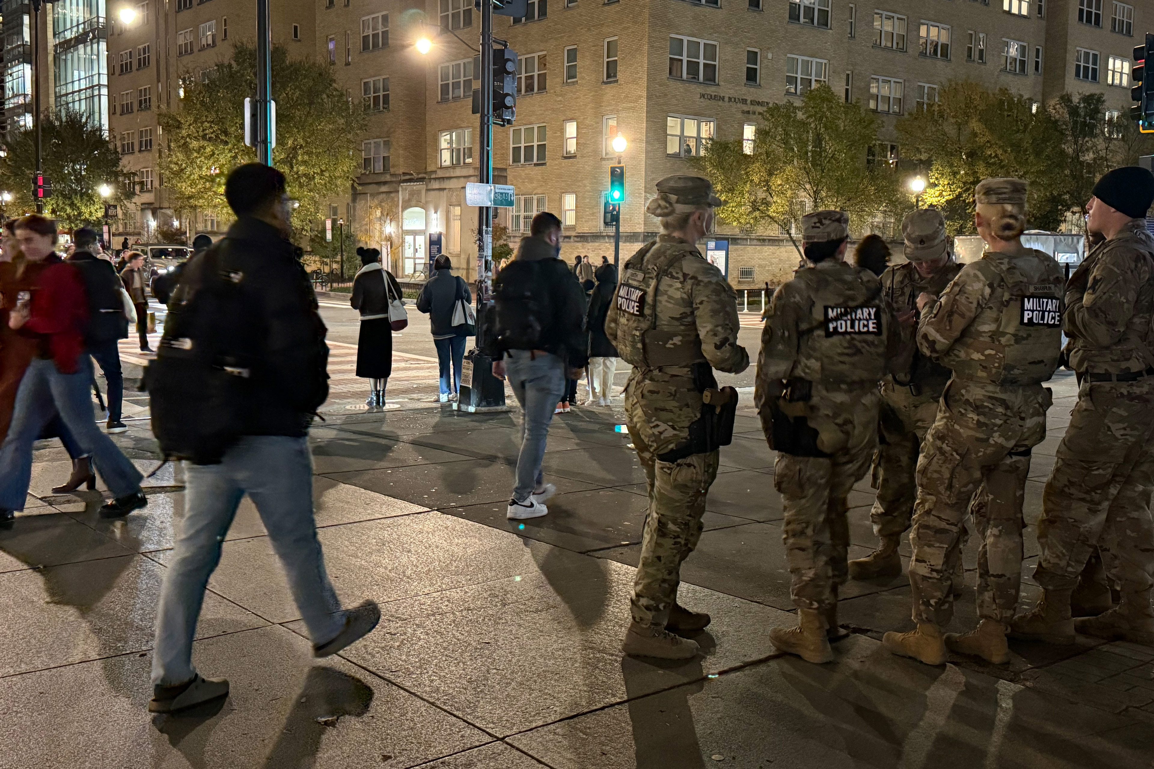 A photo of a D.C. street at night. Pedestrians walk past a set of military police officers in camouflage.