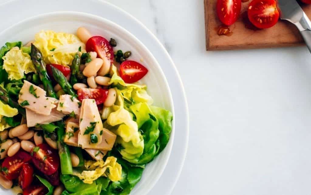 A white plate holds a mixed salad with lettuce, asparagus, tomatoes, canned beans, and chunks of tuna. Next to the plate on a wooden cutting board are halved cherry tomatoes and a knife. MyFitnessPal Blog