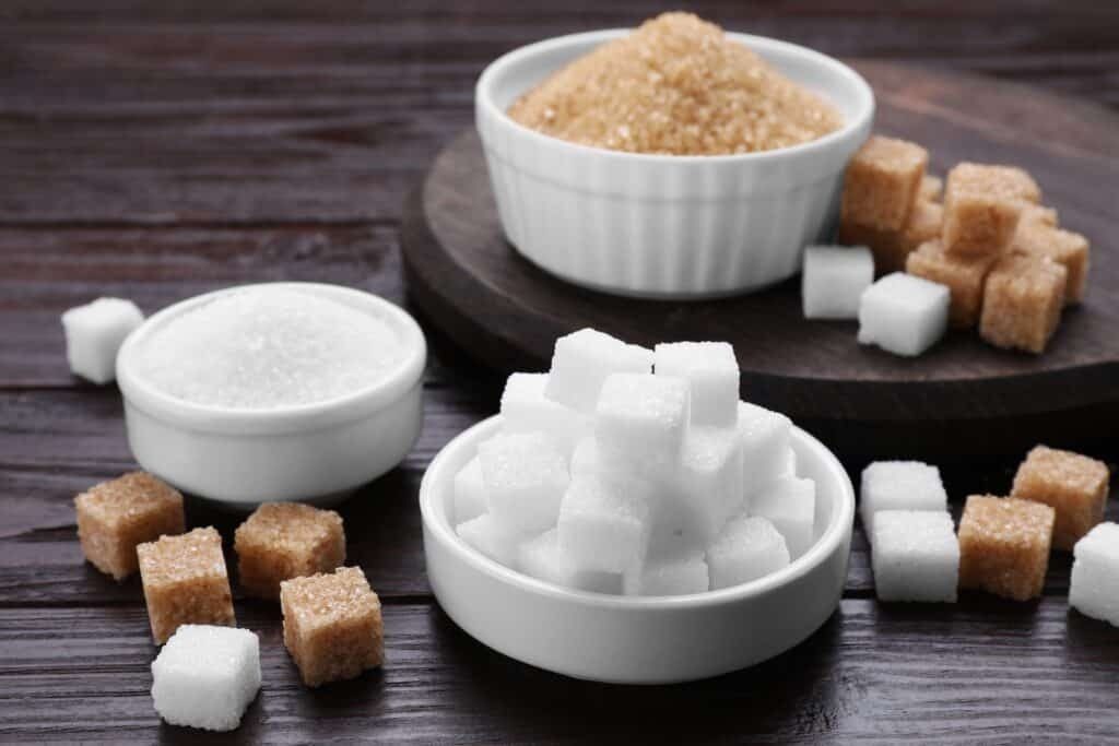 Various types of sugar in bowls including white sugar cubes, brown sugar, and granulated sugar on wooden table