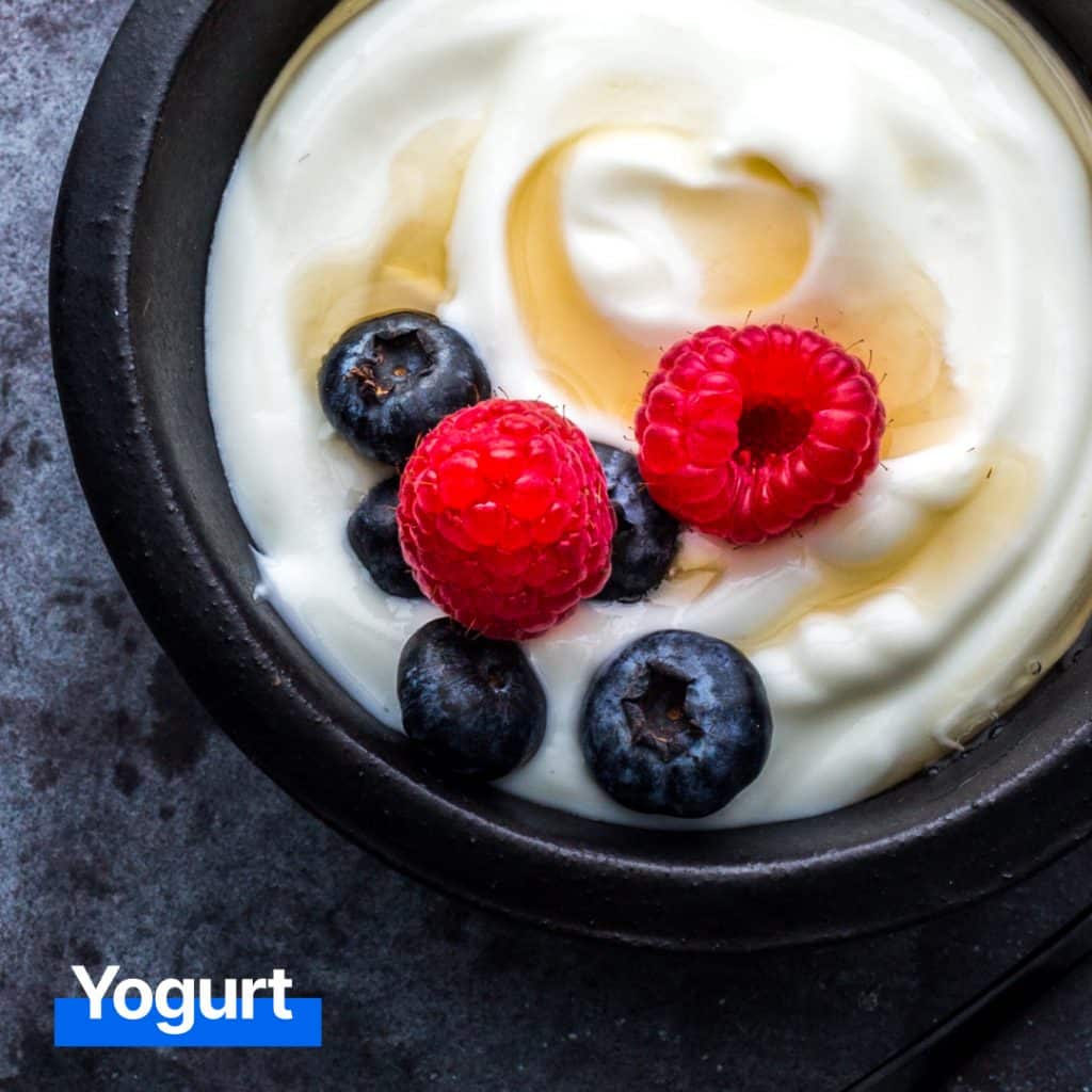 A bowl of yogurt with a smooth, creamy texture is garnished with two raspberries, three blueberries, and a drizzle of honey. Promoting foods for gut health, the word "Yogurt" is written in blue text at the bottom left. MyFitnessPal Blog