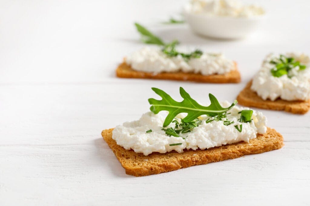 Whole grain crackers topped with cottage cheese, arugula and fresh herbs on white background