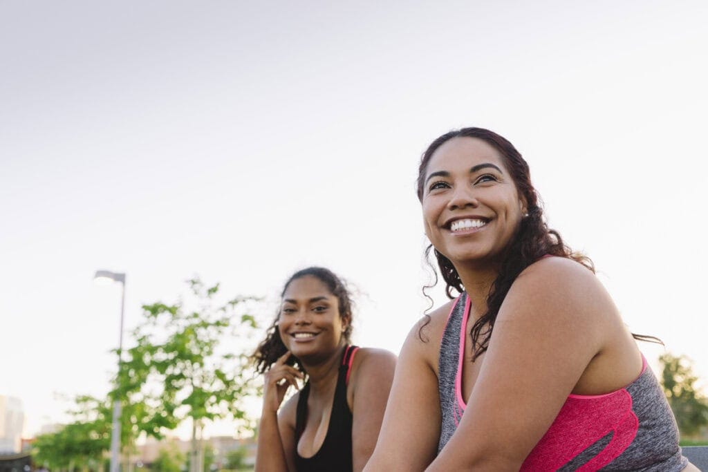 Two smiling women in athletic wear outdoors, one in pink sports bra, one in black, enjoying fitness together