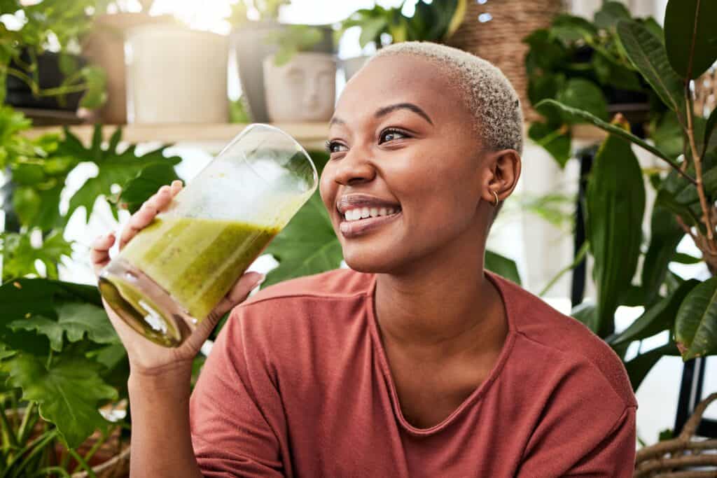 Smiling woman with short blonde hair drinking green smoothie surrounded by houseplants