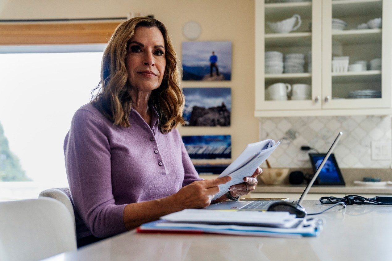 Jan Anderson sits at a kitchen island counter. A laptop and paperwork is in front of her. She holds paperwork in her hands.