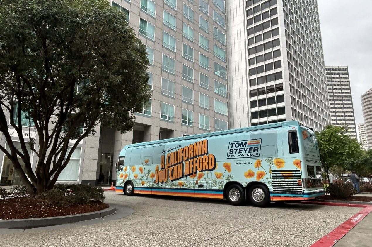 A bus with the message "All Aboard For A California You Can Afford" and "Tom Steyer for Governor" on its side is parked outside tall buildings.