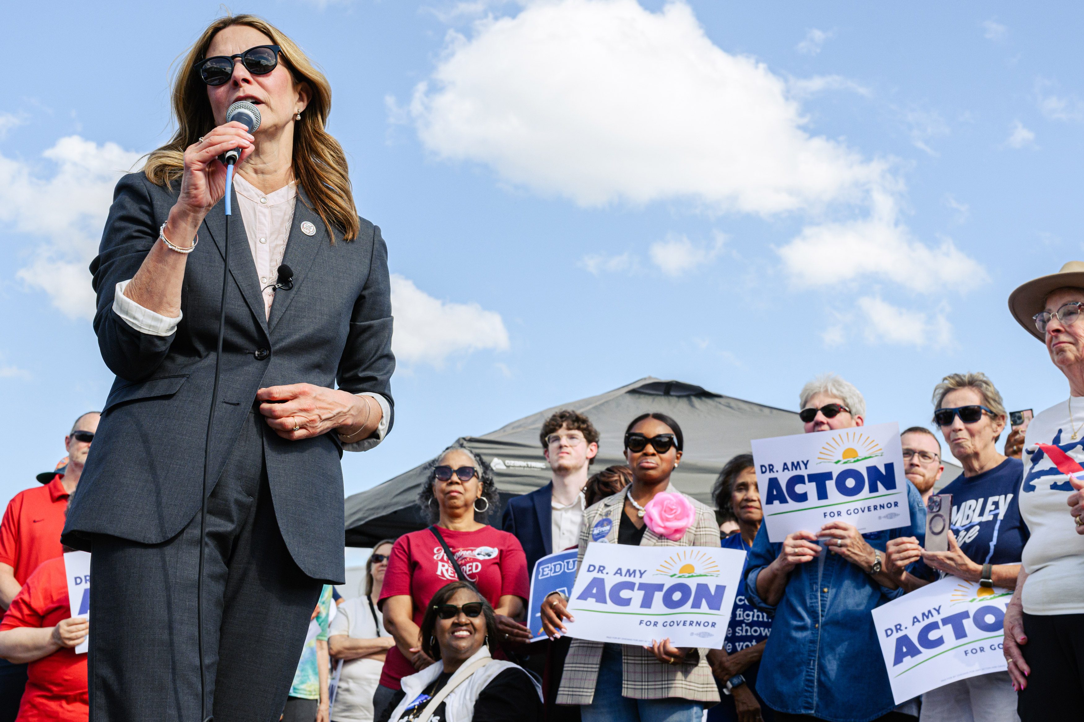Amy Acton stands, speaking to a crowd with a microphone. Around her, supporters hold signs that read, "Dr. Amy Acton for Governor."